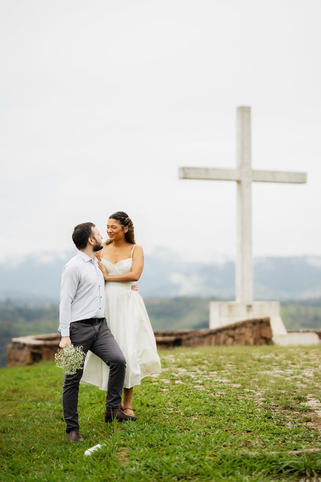 O casal se olhando de perto ao lado da cruz branca, com neblina ao fundo