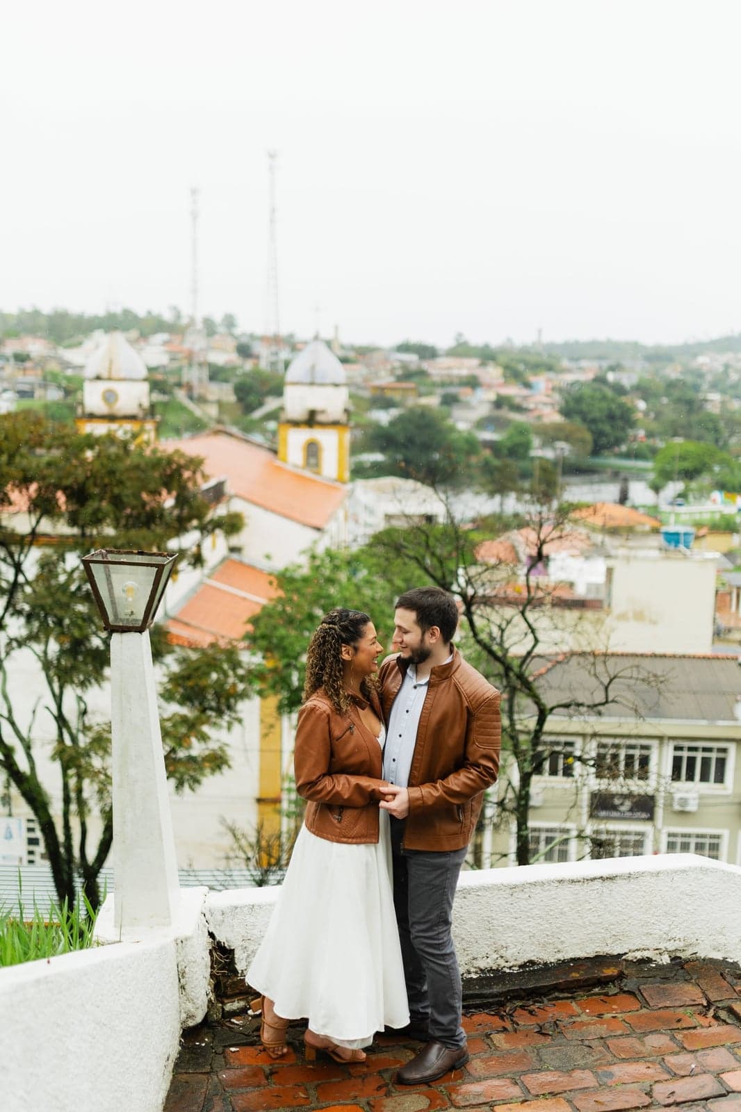 O casal caminhando abraçado por um mirante com a cidade e a igreja ao fundo
