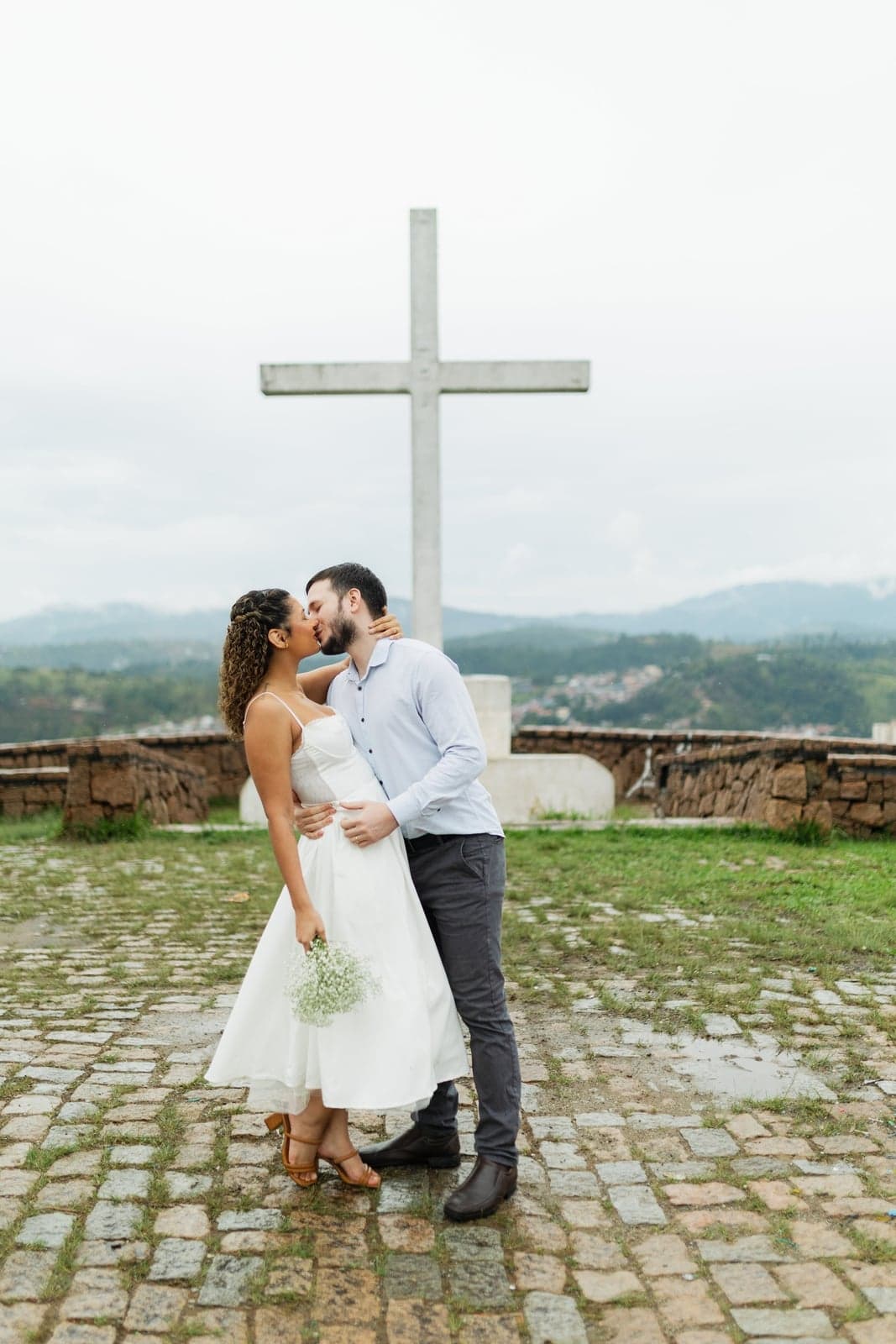 O casal se beijando ao lado da cruz branca, cercados por campo verde