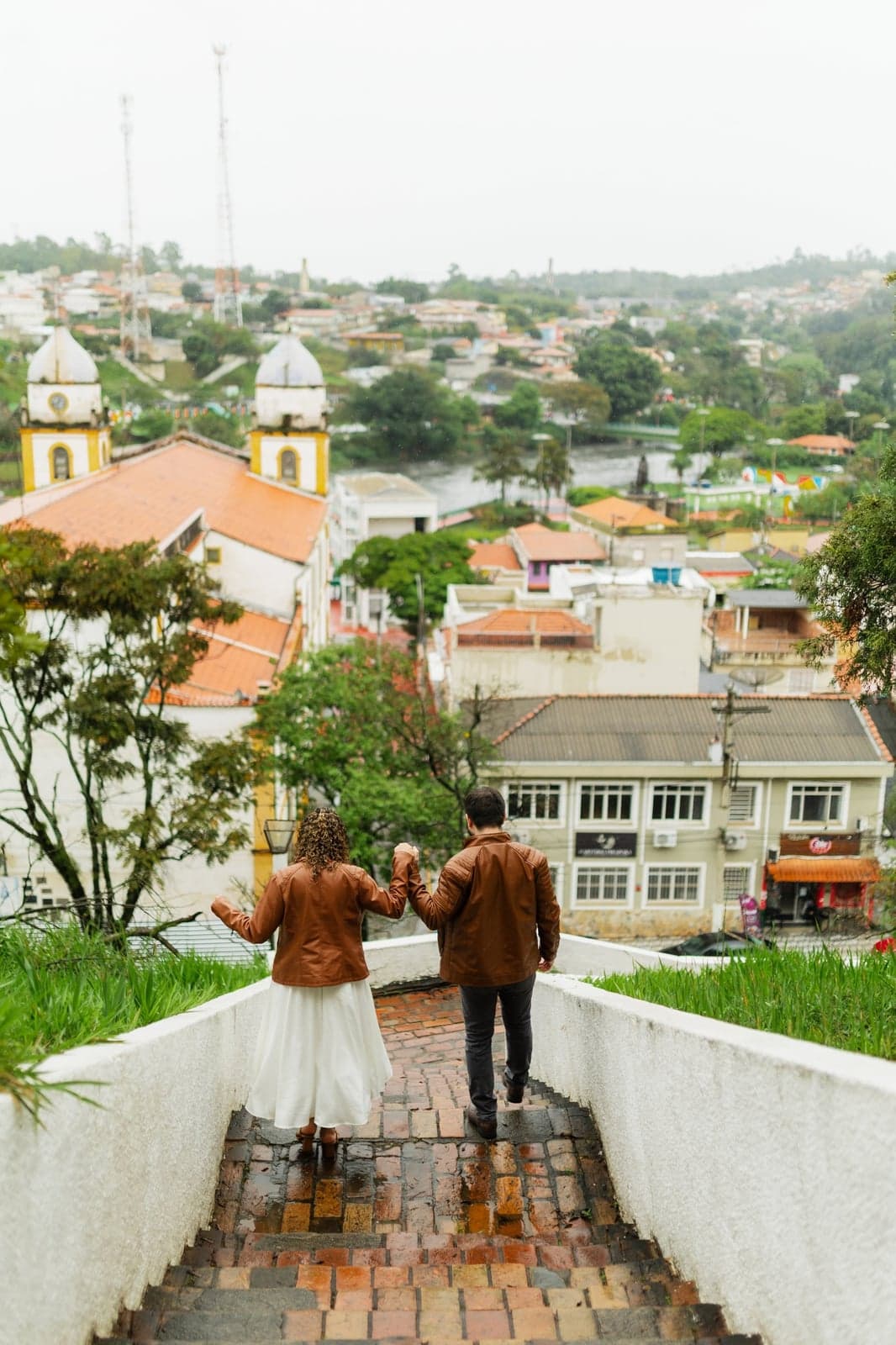 O casal caminhando de mãos dadas por uma passarela com a cidade ao fundo
