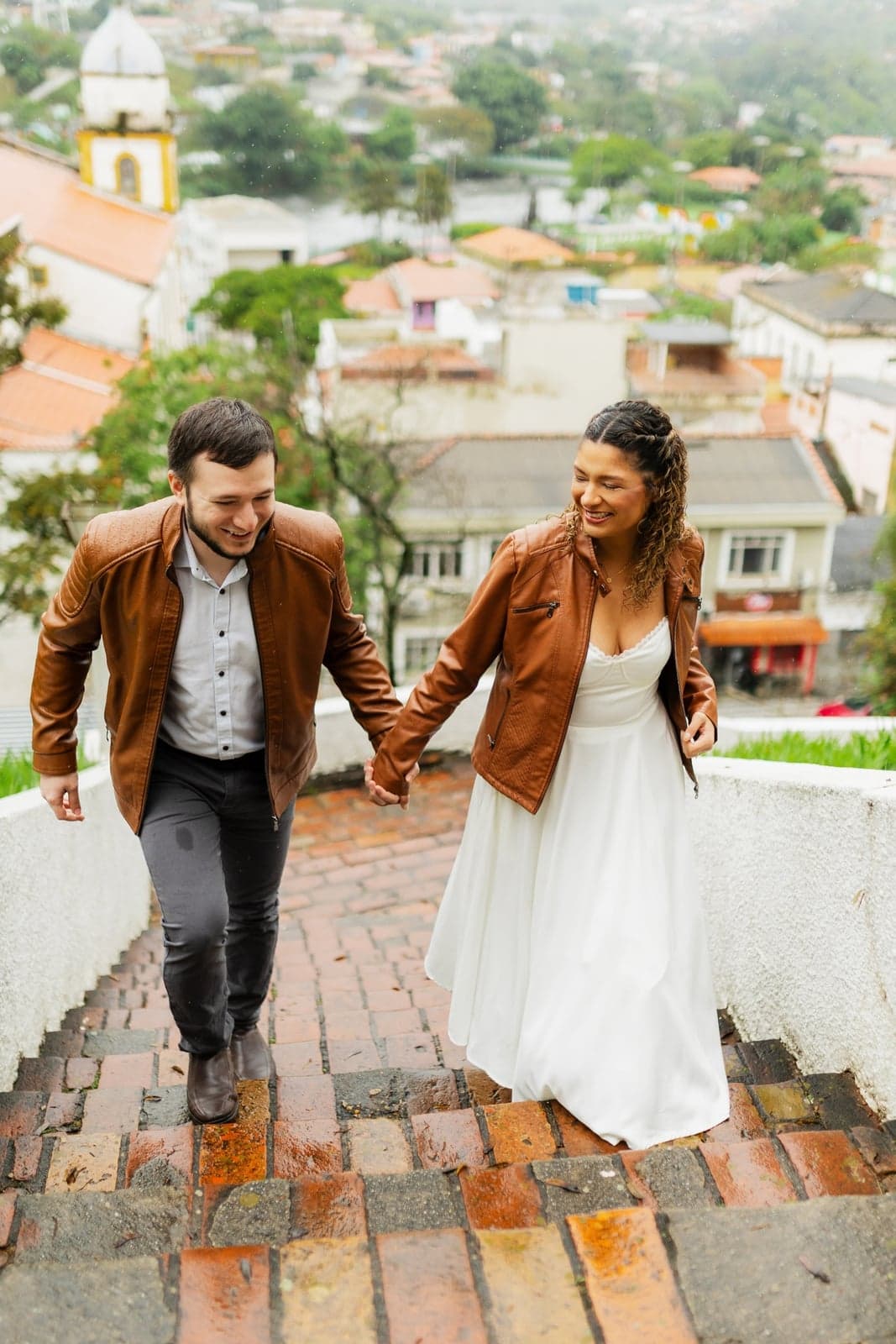 Jessyana e Pedro descendo uma escadaria de pedra de mãos dadas, com a cidade ao fundo