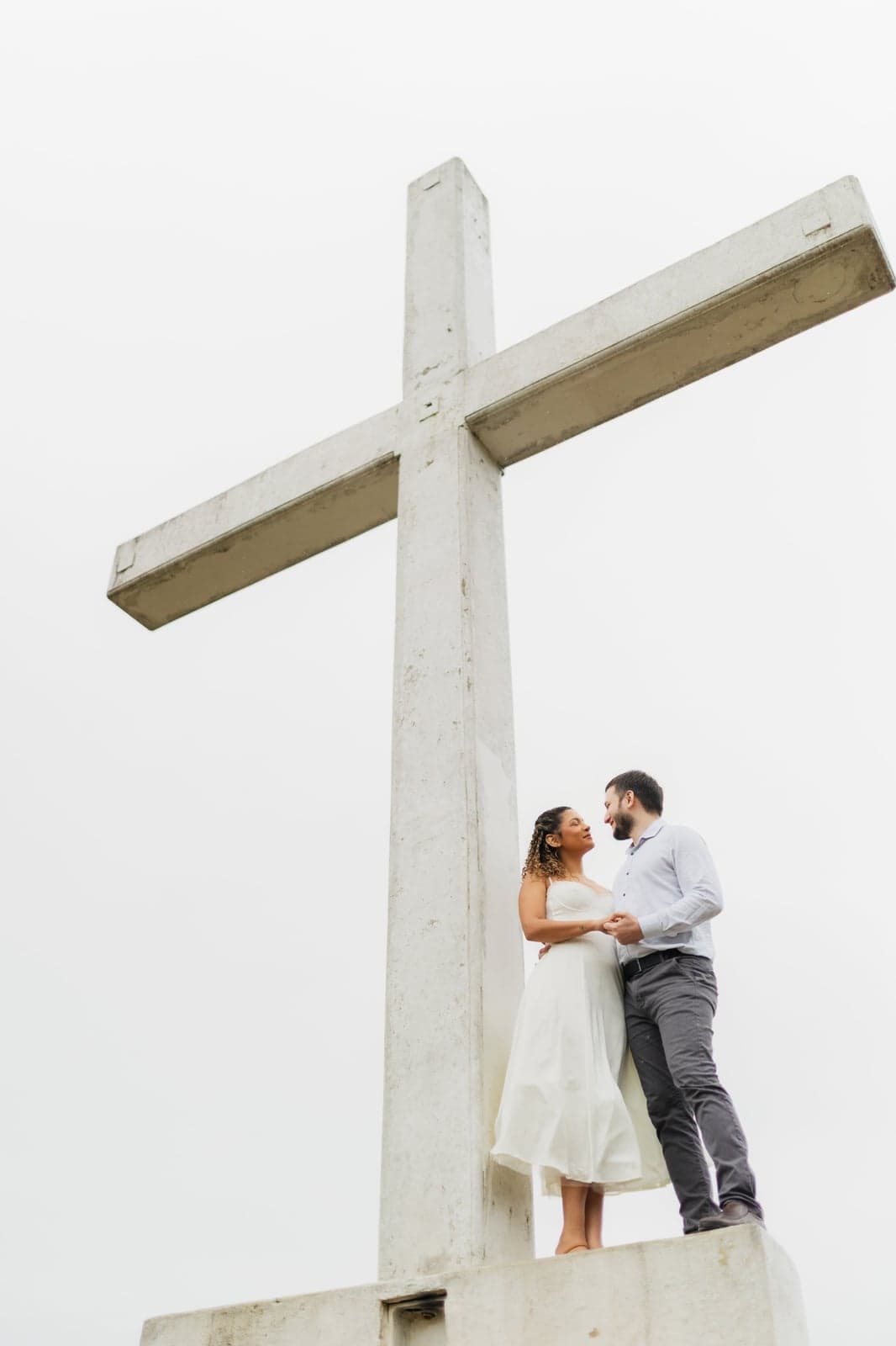 Jessyana e Pedro se olhando abraçados ao lado de uma cruz branca, com neblina nas montanhas ao fundo